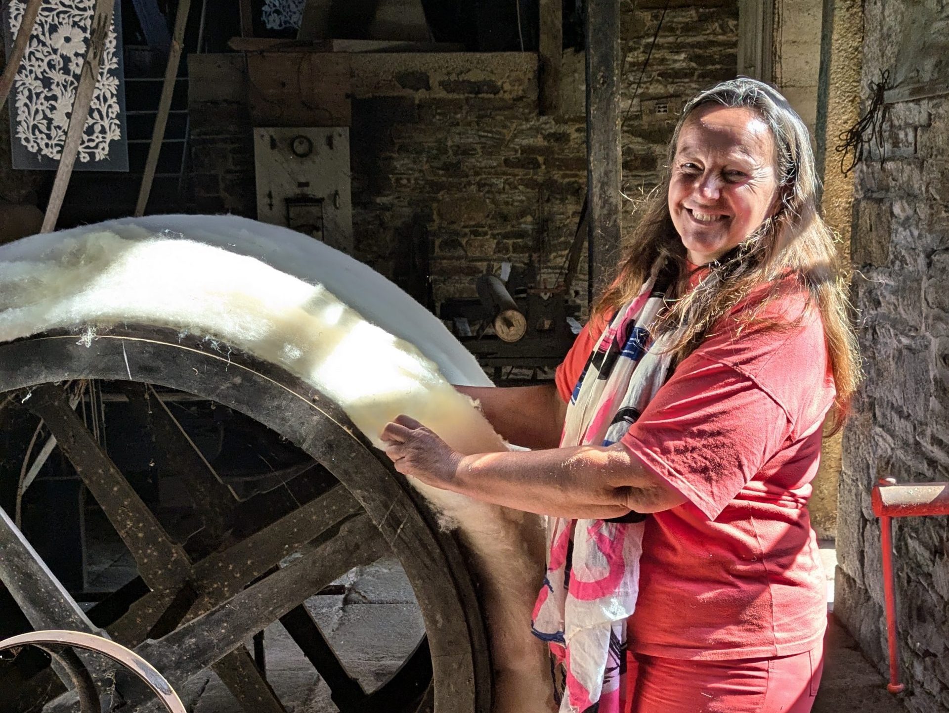 Une femme en chemise rouge se tient à côté d'une grande roue du moulin de la filature, manipulant une couche de laine brute dans un atelier rustique éclairé par le soleil.